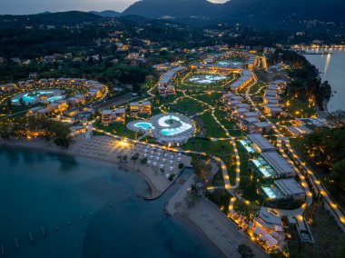 Bir hava görüntüsü, kıyı şeridinde çok sayıda bina, havuz, plaj ve alacakaranlıkta verimli peyzaj içeren iyi ışıklandırılmış bir tatil beldesini ortaya çıkarıyor..