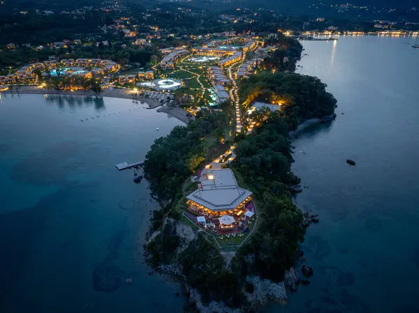 Bu hava çekimi, alacakaranlıkta bir sahil beldesini yakalıyor. Aydınlatılmış binalar ve yarımadadan geçen bir patika var..