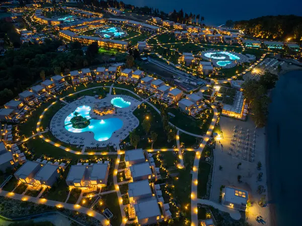 Yukarıdan çekilen gece sahnesinde bir tatil köyü, aydınlatılmış yollar ve deniz tarafından yerleştirilmiş havuzlar büyüleyici bir ortam yaratıyor..