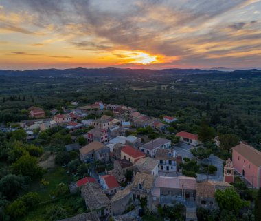 Gün batımında gökyüzüne karşı çerçevelenmiş güzel bir köy. Yuvarlanan tepeler ve ufukta yoğun ormanlar var. Kırmızı çatılı binalar göze çarpıyor.