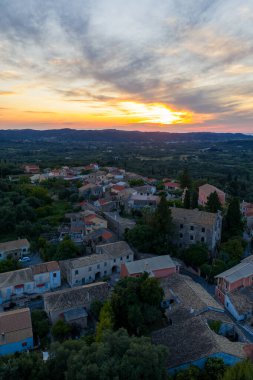 Manzaralı bir hava manzarası, güneş batarken ağaçlarla çevrili küçük bir köyü yakalar. Gökyüzünü sarı ve turuncu renklerle boyarlar..
