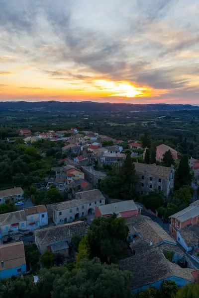 Manzaralı bir hava manzarası, güneş batarken ağaçlarla çevrili küçük bir köyü yakalar. Gökyüzünü sarı ve turuncu renklerle boyarlar..