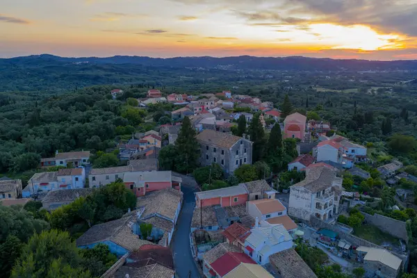 Havadan çekilen görüntülerde çeşitli renkli binalar ve yemyeşil çatılarla çevrili kırmızı fayanslı bir köy görülüyor. Altın bir günbatımı ufku süslüyor.