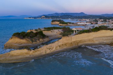 Havadan bakıldığında sakin bir deniz manzarasında kıyı şeridi boyunca katmanlı kaya oluşumları görülüyor. İnsanlar gün batımı manzaralı kayalarda toplanır..