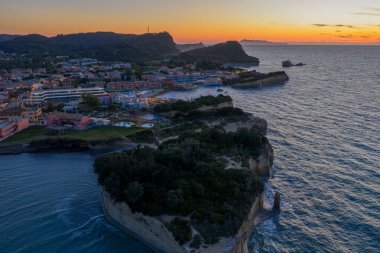 Sahilde bir kıyı kasabası, bir uçurum ve deniz manzarası altın ışıkla ışıldayan canlı bir günbatımında. Yeşil doğa ve nazik dalgalar manzarayı tamamlar..