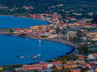 Alacakaranlıkta canlı bir sahil kasabasının manzarası yemyeşil bir yamaçla sakin suların arasına kurulmuş renkli binalar sergiliyor..