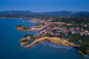 Kıyı yerleşiminin havadan görünüşü eşsiz kaya oluşumlarıyla karanlık bir gökyüzünün altında denizle buluşuyor, uzak dağların arka planına kuruluyor..