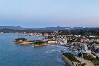 Sahil boyunca uzanan pitoresk bir kasaba, denizle buluşan eşsiz kaya oluşumlarına ev sahipliği yapıyor. Arkaplanda renkli bir alacakaranlık altında dağlar yükselir..