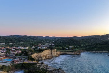 Sakin bir sahil kasabası yemyeşil tepeler ve çarpan kayalıklarla çevrelenmiş. Gökyüzü alacakaranlıkta gün batımının yumuşak renkleriyle boyanmış..