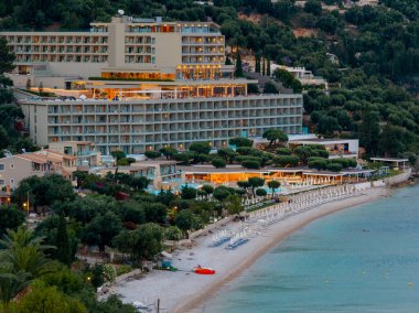 Turkuaz su ve çevreleyen tepeleri çevreleyen yeşil ağaçların yanında, beyaz şemsiyeli büyük lüks bir tatil köyü bir plajda yer alır..