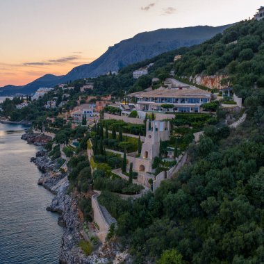 Denize dökülen teras bahçeleri ile bir uçurumun üzerinde büyük bir villa yer almaktadır. Dağlar alacakaranlıkta yükselir..
