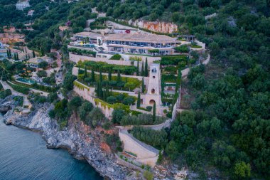 Bu resim, mimariyle doğayı harmanlayan bir uçurumun kenarındaki geniş bir malikaneyi gösteriyor. Peyzajlı bahçeler ve okyanus manzarası dingin bir zemin oluşturur.