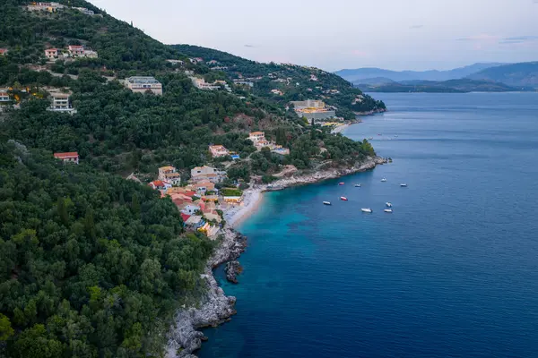 Hava manzarası, yeşil bir yamaçta evleri yakalar ve alacakaranlıkta sakin, derin bir mavi denizde teknelerle kumsalla karşılaşır..