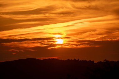 Altın saat gün batımı, dağlık bir arazide alevli bir ışık saçıyor. Gökyüzünde bulutlar dolaşıyor, derinlik ve dram katıyor..