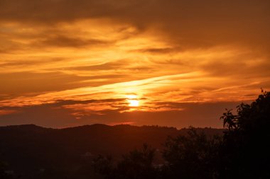 Huşu uyandıran bir günbatımı, portakal ve altının canlı tonlarıyla gökyüzünü boyar. Yuvarlanan tepeler ve ağaçlar resim gibi bir siluet oluşturuyor..