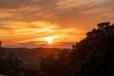 Canlı turuncu bir gün batımı, ağaçlarla dolu, tepecikli bir manzarayı aydınlatır. Sıcak ve yumuşak renklerle sakin ve pitoresk bir akşam sahnesi yaratır..