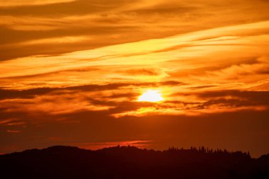 Canlı bir altın gün batımı, sıcak renklerde parlayan bulutlarla gökyüzüne hükmediyor. Ağaçlardan ve tepelerden oluşan bir siluet sakin bir ufuk oluşturur..