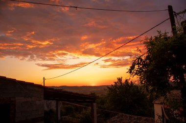 Renkli bir günbatımı, tepelerin, ağaçların, elektrik hatlarının siluetlerinin ve manzarayı noktalayan yapı elementlerinin olduğu kırsal bir arazide patlıyor..