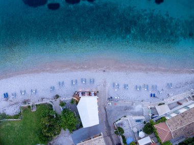 Bu kuş bakışı manzara, kristal berrak turkuaz sularıyla, şezlonglarla ve bitişik tatil yerleriyle çevrili güzel bir çakıl taşı plajını gösteriyor..