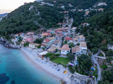 Bu resim yeşil tepelerin kucakladığı canlı bir köyü tasvir ediyor. Güzel bir çakıl taşı plajına ve turkuaz denize dökülüyor. Huzurlu bir sahil inzivası sunuyor..