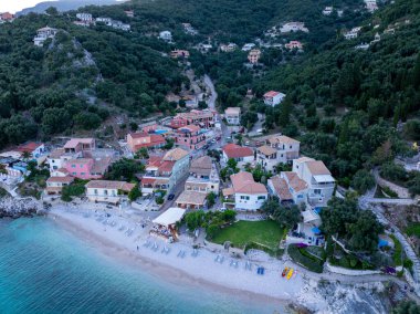 Deniz kenarındaki manzaralı bir köyde, yoğun ağaçlarla kaplı bir yamaçla desteklenen şezlonglu plaj boyunca renkli binalar bulunur..