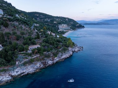Deniz manzaralı ağaçlar ve evlerle kaplı bir tepe ile çarpıcı bir kıyı şeridini gösteren hava görüntüleri. Bir tekneyle berrak mavi sular.