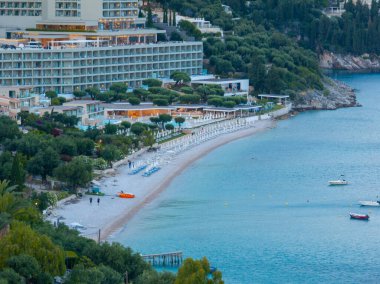 Sahil manzaralı sakin bir sahil, kumlu bir sahil, temiz su ve çevresini saran yemyeşil ağaçlar. Tekneler denizde yüzüyor..