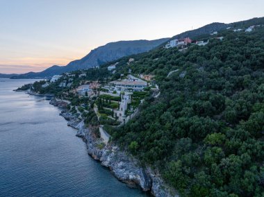 Hava manzarası, alacakaranlıkta Hırvatistan 'ın dağlık bir kıyısında solunda deniz bulunan ağaçlarla çevrili bir uçurum kenarındaki binaları gösteriyor..
