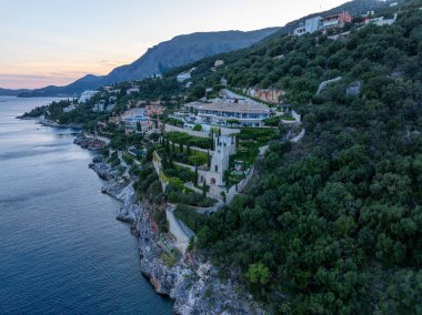 Bu manzarada ağaçlarla kaplı bir yamaçta inşa edilmiş yapılar kayalık kıyı şeridine ve aşağıdaki mavi denize dökülüyor..