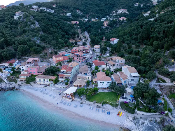 Deniz kenarındaki manzaralı bir köyde, yoğun ağaçlarla kaplı bir yamaçla desteklenen şezlonglu plaj boyunca renkli binalar bulunur..