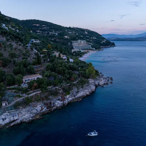 Yamaçta yerleşik binaların bulunduğu bir sahil manzarası, alacakaranlıkta sakin bir denizle karşılaşma, suda bir tekne..