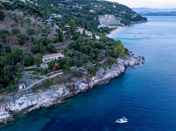 Bir hava görüntüsü kayalık bir sahil manzarası, bol ağaçlar, yamaçtan evler ve büyük bir tatil köyü olan kumlu bir sahil görüntüsü yakalar.. 