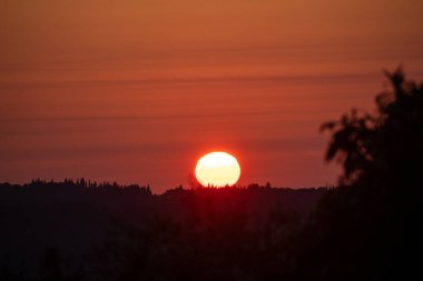 Canlı bir günbatımı, güneş ufuktan aşağı batarken ve ağaçları siluetlerken ve sakin bir ortam yaratırken sıcacık renklerle bir manzarayı yıkıyor..