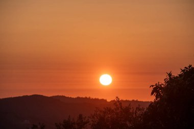 Canlı bir günbatımı, turuncu ve kırmızı tonlarda yüzen yuvarlanan tepeler ve siluetli ağaçlardan oluşan bir manzara sergiliyor..