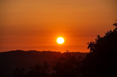 Huzurlu bir sahne, tepelik bir arazide canlı bir gün batımı sergiler. Gökyüzü turuncu ve kırmızı tonlarıyla parlıyor, huzurlu bir manzara yaratıyor..
