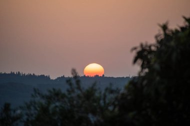 Alacakaranlıkta yakalanan güneş, siluetli ağaçlarla çevrili uzak bir ufukta zarifçe batar ve manzaraya turuncu bir parıltı yayar..