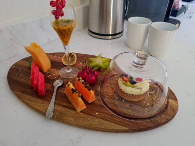 Çeşitli taze meyvelerle dolu ahşap bir tepsi, bir bardak tutku meyvesi suyu, ve cam bir örtünün altında küçük bir tatlı, zarif bir şekilde tezgahın üzerine yerleştirilmiş..