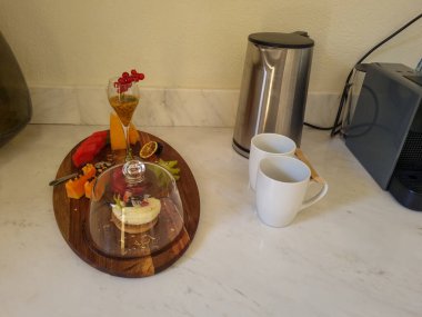 Zarif bir kahvaltı düzeninin yakın çekimi: tropikal meyve, hamur işi, meyve suyu, kahve makinesi ve mermer üzerinde kupalar. Lüks bir otel havası..