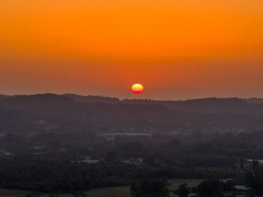 Turuncu bir günbatımı aşağıdaki manzaraya bir parıltı saçar, siluetli tepeler yukarıdaki canlı gökyüzüne bir zemin sağlar.. 