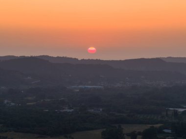 Yüksek bir bakış açısından yakalanan bu görüntü, güneşin gökyüzünü renklerle boyadığı kayan tepelerden ve uzak yapılardan oluşan bir manzara sunuyor..