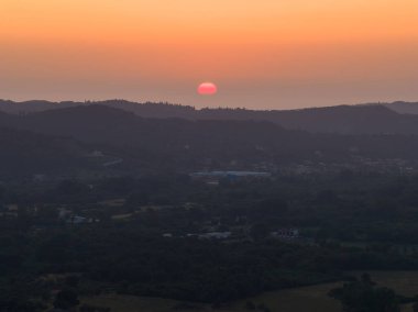 Yuvarlanan tepelerin üzerinde büyüleyici bir günbatımı, gökyüzüne turuncu bir parıltı saçıyor. Uzak köy huzurlu manzaraya derinlik katıyor..