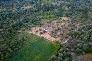 Yeşillik ağaçlar, bitki örtüsü, toprak yolları ve kırsal kesimdeki bitişik bir araziyle çevrili bir binayı gösteren bir hava perspektifi..