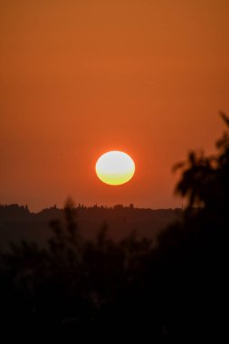 Canlı bir günbatımı ufukta turuncu bir parıltı bırakır, uzak tepeleri ve ağaçları gölgeler. Doğanın nefes kesici gün sonu manzarası..