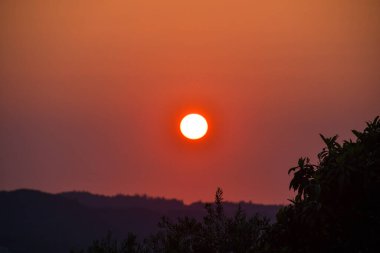 Ufukta parlayan güneş, turuncu ve kırmızı tonlarla gökyüzünü aydınlatıyor, dingin bir sahne için tepeleri ve ağaçları siluetliyor..