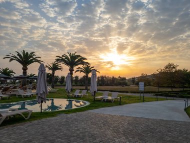 Palmiye ağaçlarının siluetleriyle dolu sakin bir tatil köyü havuzu üzerinde nefes kesici bir gün doğumu. Güneş ışınları dramatik bulutlarda parlıyor. Mükemmel bir tropikal tatil için güzel bir gün vadediyor.