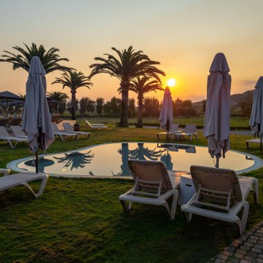 Lüks bir tatil köyünde sakin bir gün doğumu, ufukta altın güneş yükseliyor. Palmiye ağaçları berrak bir gökyüzüne karşı siluetlenir, sakin bir havuza yansıtılır, mükemmel bir tatil ortamı yaratır.