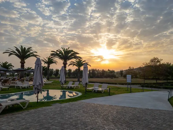 Palmiye ağaçlarının siluetleriyle dolu sakin bir tatil köyü havuzu üzerinde nefes kesici bir gün doğumu. Güneş ışınları dramatik bulutlarda parlıyor. Mükemmel bir tropikal tatil için güzel bir gün vadediyor.