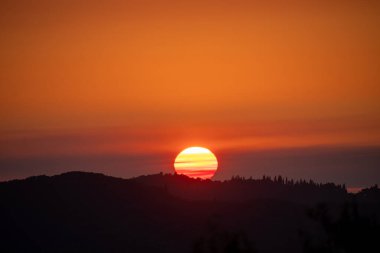 Batan güneşin ışıldayan küresi, karanlık tepelerin üzerinde sisli, sıcak turuncu ve kırmızı bir gökyüzü olan ağaç çizgili bir sırtın siluetinin arkasında kaybolur.