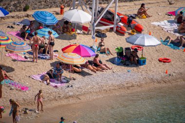 Turistlerin şemsiye altında güneşlendiği, çocukların güneşli bir günde suyun kenarında oynadığı hareketli bir çakıl taşı plajının yüksek açılı görüntüsü.