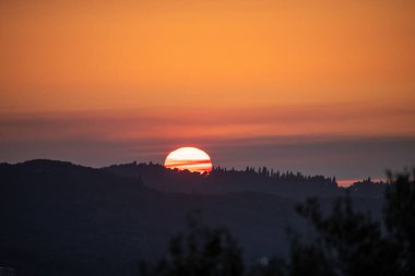 Batan güneşin ışıldayan küresi, karanlık tepelerin üzerinde sisli, sıcak turuncu ve kırmızı bir gökyüzü olan ağaç çizgili bir sırtın siluetinin arkasında kaybolur.